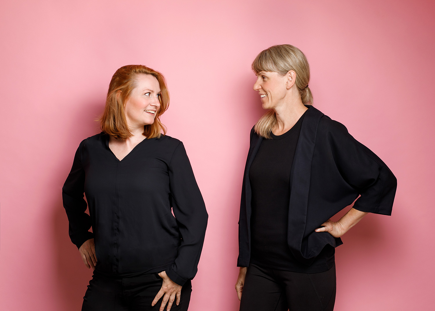 Ein Foto von Katy Schmitt und Jule Demel vor einem pinkfarbenem Hintergrund. Katy steht links und dreht sich zu Jule um. Jule steht rechts und stützt einen Arm in die Seite. Beide tragen schwarze Kleidung und lachen.