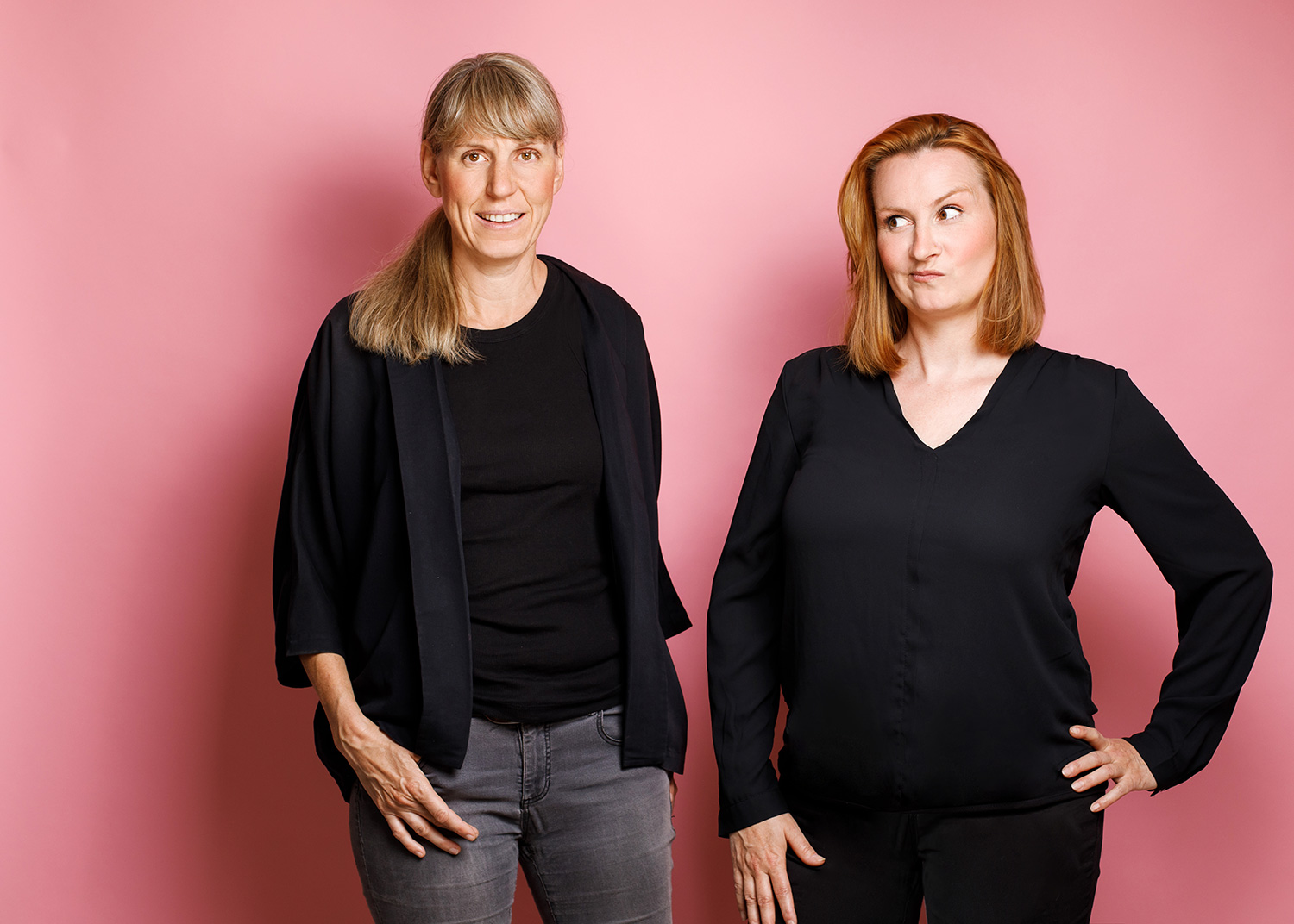 Ein Foto von Jule Demel und Katy Schmitt vor einem pinkfarbenem Hintergrund. Jule steht links und steckt eine Hand in die Hosentasche. Katy steht rechts macht ein lustiges Gesicht. Beide tragen schwarze Kleidung.