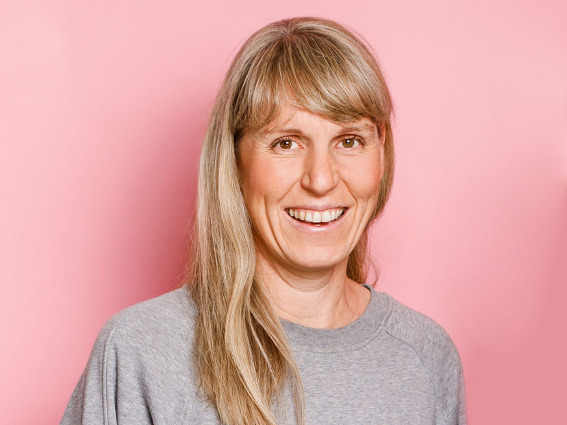 jule-demel-grafikdesign-dresden Ein Portraitfoto von Jule Demel vor pinkfarbenem Hintergrund. Jule hat blonde lange Haare, lacht und blickt frontal in die Kamera.