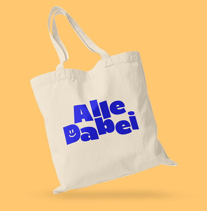 Weiße Umhängetasche mit dem royalblauen Logo von AlleDabei auf gelbem Untergrund. Das Logo ist eine schlichte, zweizeilige Wortmarke mit einem Smiley im D.
